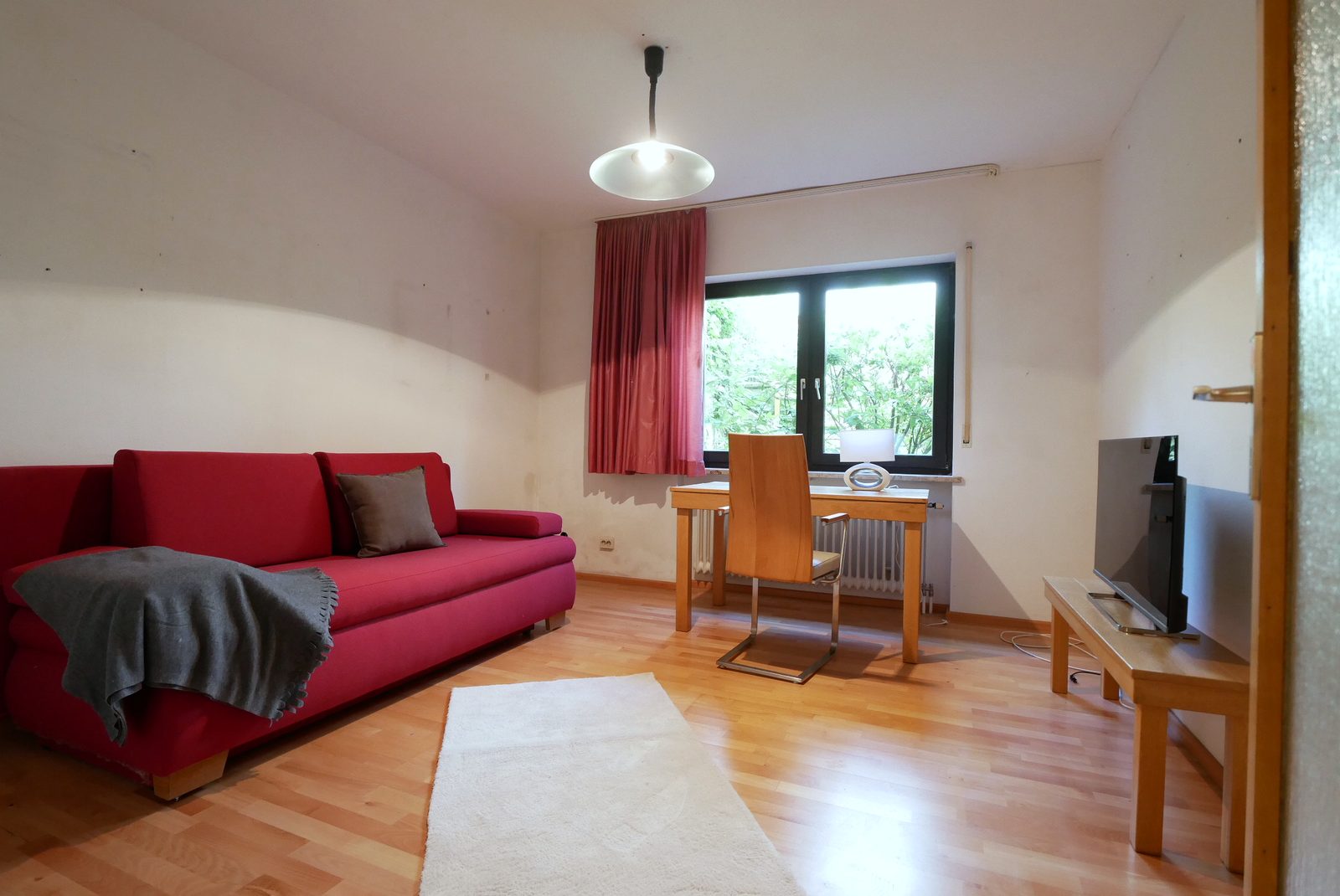 Arbeitszimmer nach dem Home Staging: rotes Sofa mit Decke, Schreibtisch am Fenster, Teppich und Fernseher