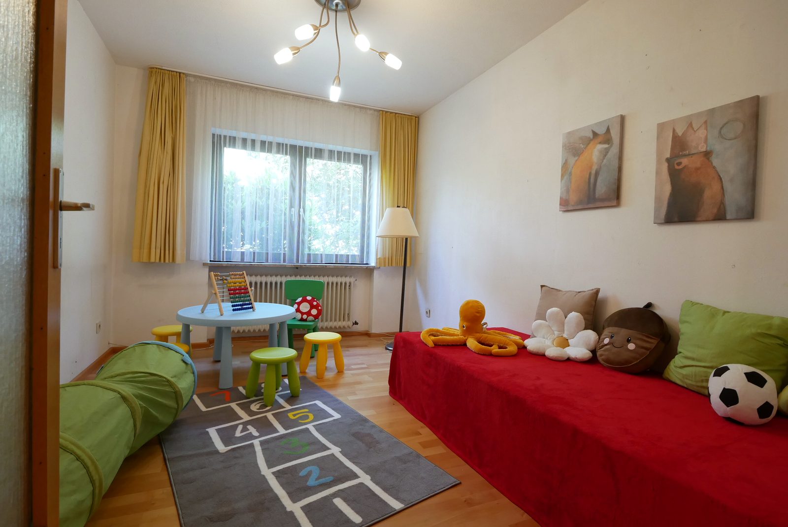 Kinderzimmer nach dem Home Staging: rotes Bett mit Kuscheltieren, Spieltisch, Hüpfkästchen-Teppich und Bilder
